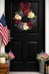 Decorative wreath with red, white, and blue flowers on a black door with American flags and plants.