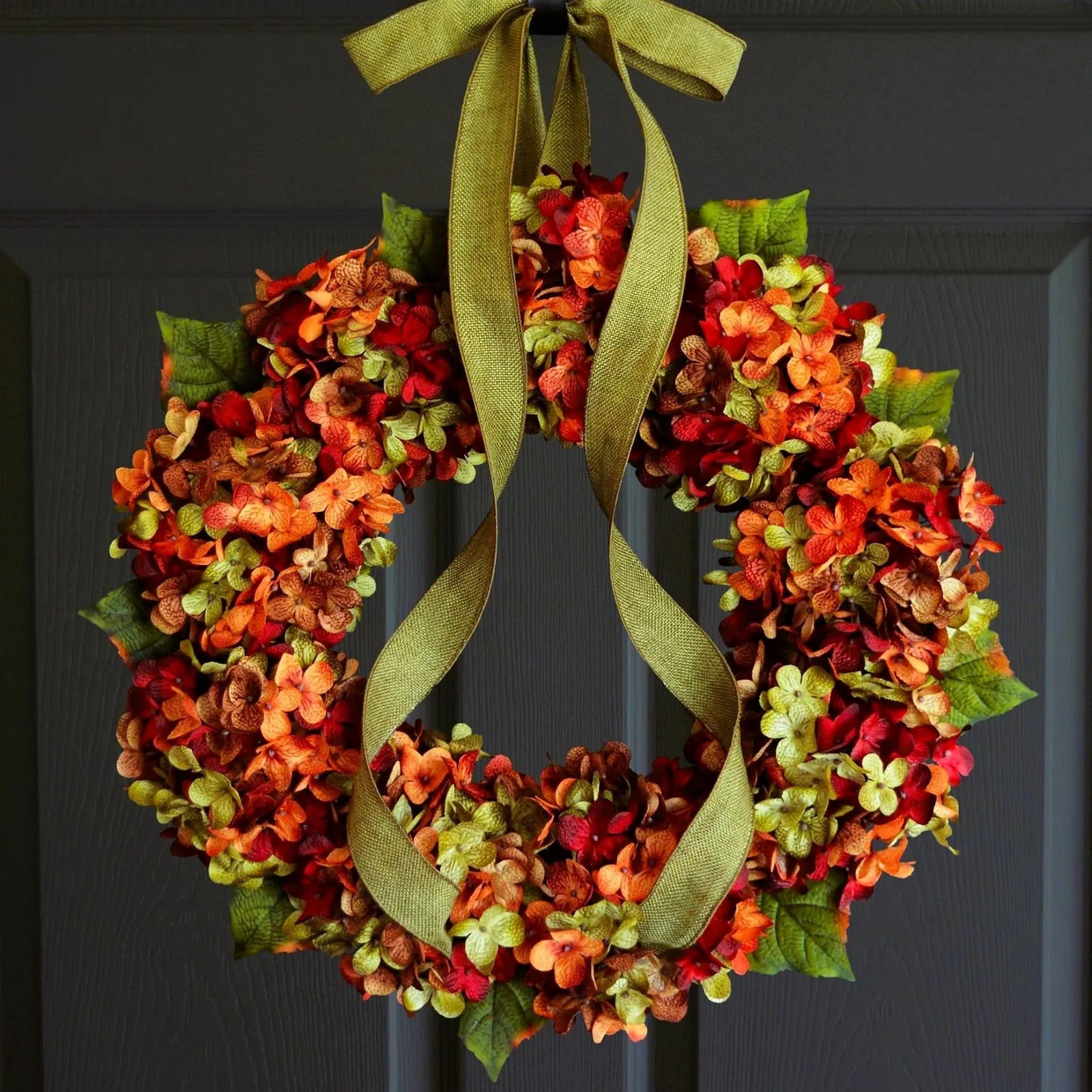 An autumn hydrangea wreath hanging on a dark gray door. The wreath features clusters of red, orange, and green hydrangea blossoms with a wide olive green ribbon tied in a bow at the top.