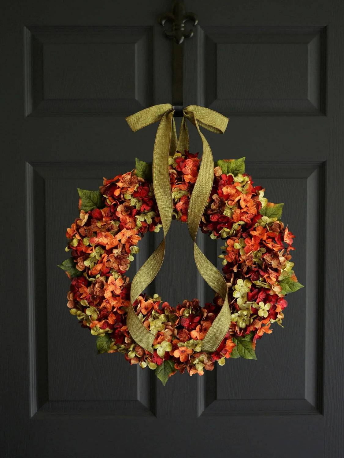 An autumn-themed floral wreath with orange, red, and yellow hydrangea-like blooms, green leaves, and an olive-green ribbon bow, hanging on a dark charcoal paneled door.